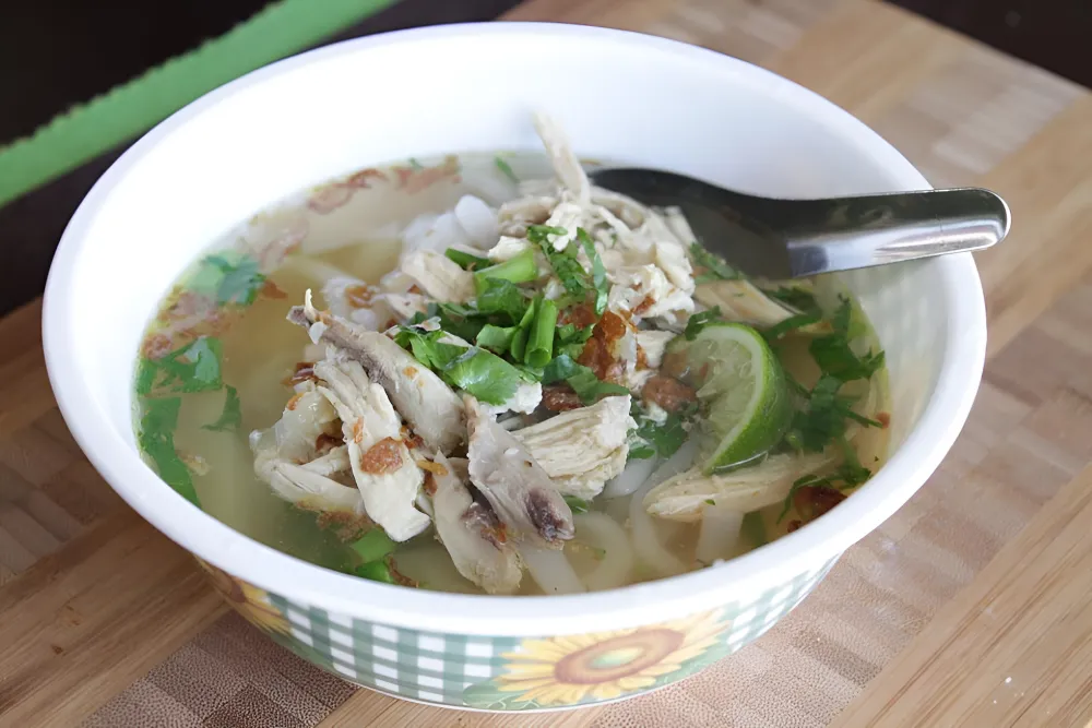 steaming hot bowl of Khao Piak Sen Laotian wet noodle soup with thick rice noodles, chicken, and fresh garnishes for breakfast